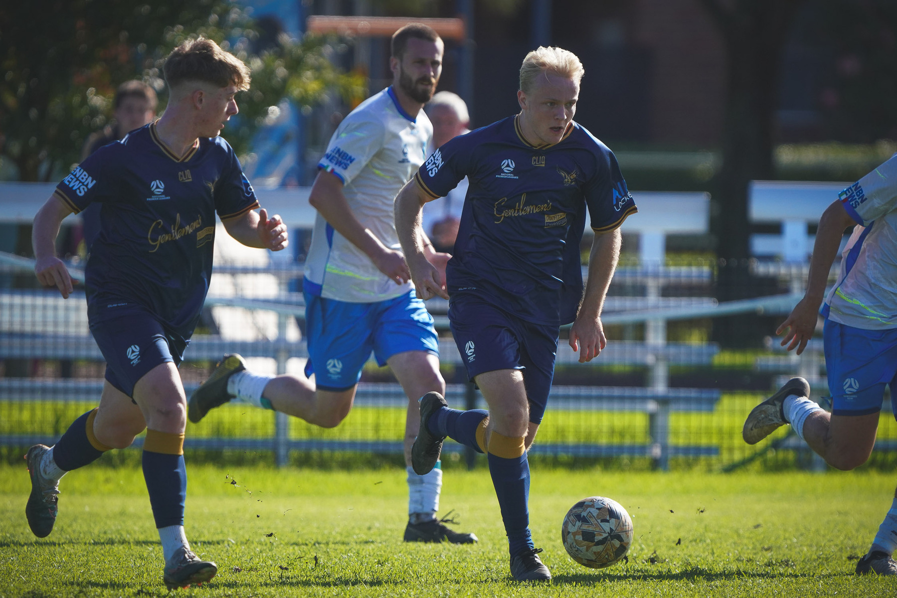 Football players in action on a sunny day, with one in a dark blue uniform controlling the ball.