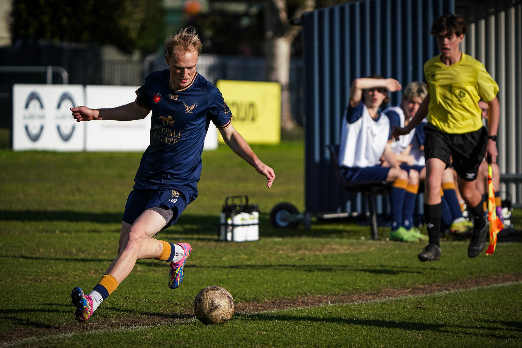 A soccer player in a navy blue uniform is kicking a ball on a grassy field while teammates and a referee watch from the sidelines.