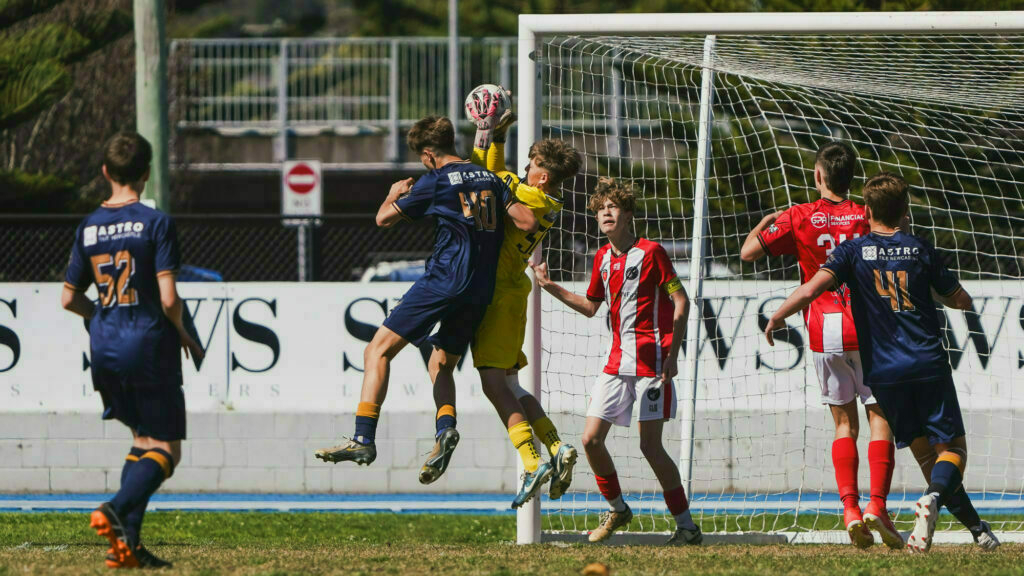 Two soccer players in blue and a goalkeeper in yellow contest for the ball in front of the goal, while other players observe the action.