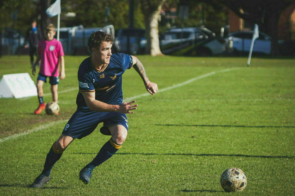 A soccer player in blue prepares to kick the ball on a grassy field, with another player in pink watching nearby.