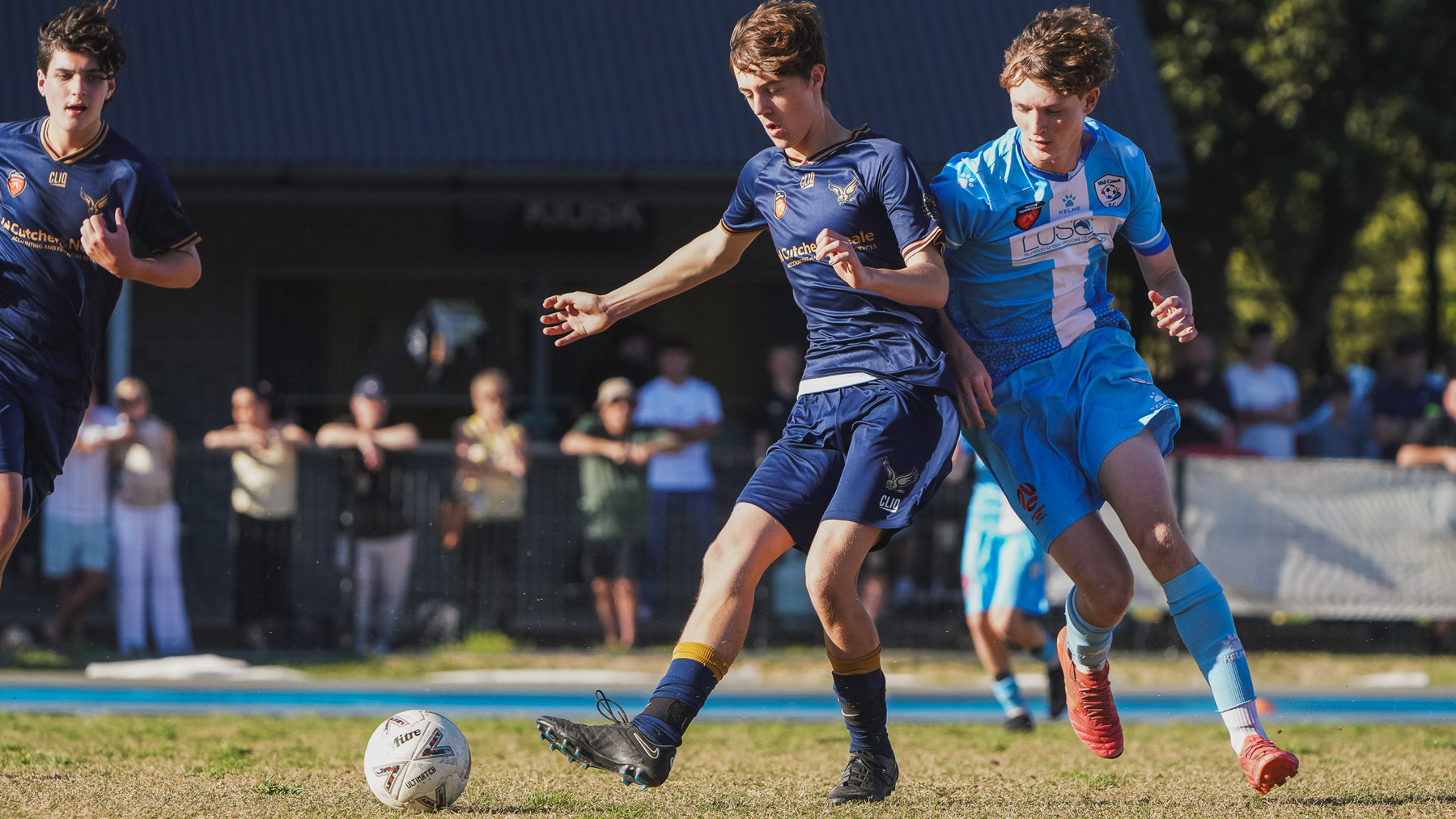 Two soccer players are competing for the ball during a match, with spectators watching in the background.