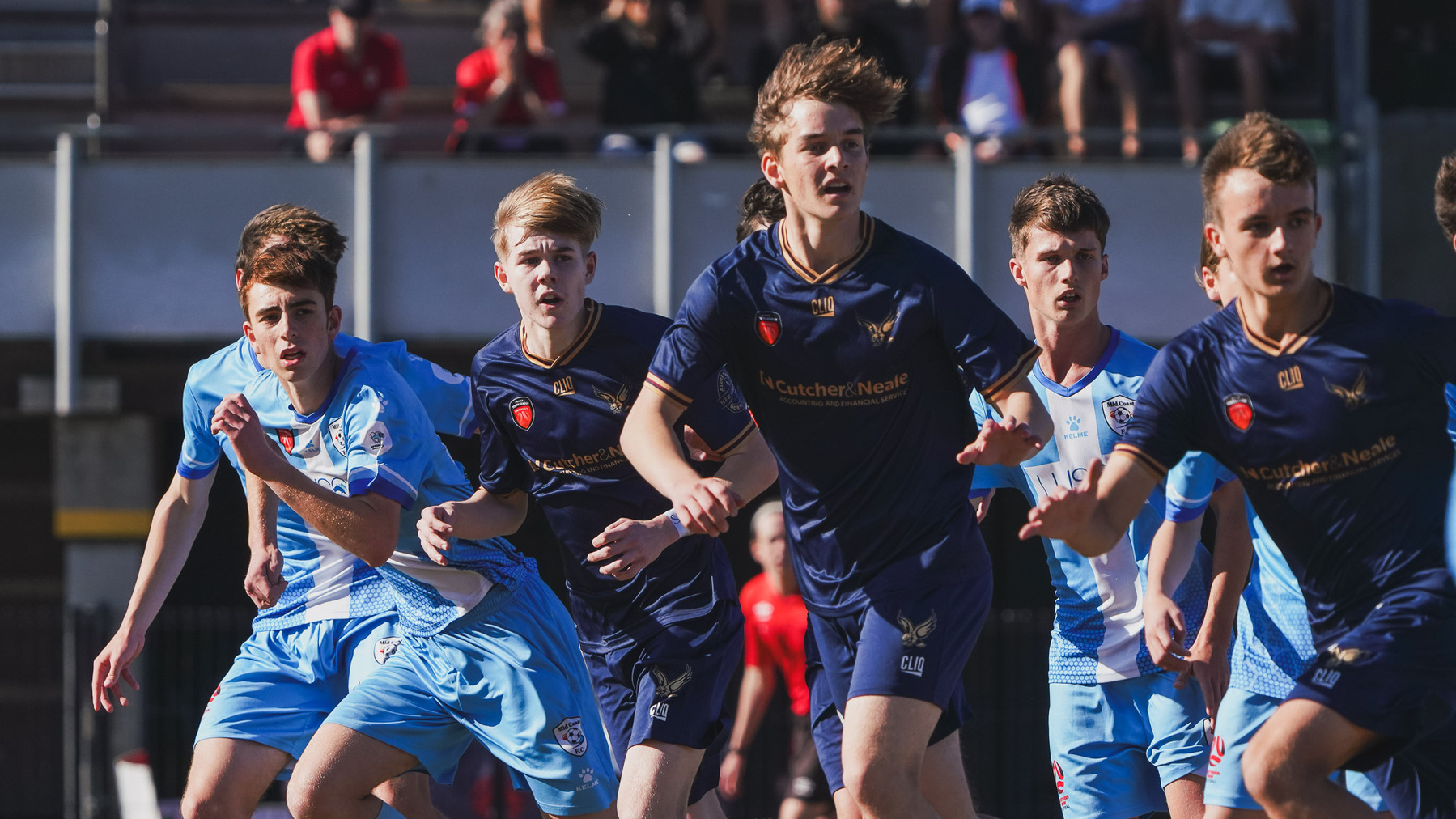 A group of young soccer players in blue uniforms is actively engaged in a game, with a focused crowd in the background.