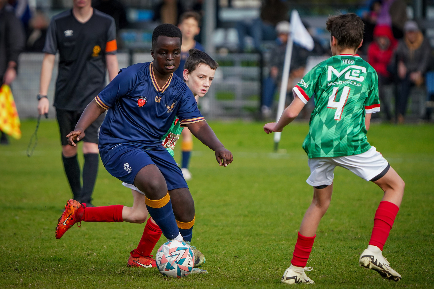 A youth soccer match is in progress with one player in blue dribbling the ball while being challenged by opponents in green jerseys.