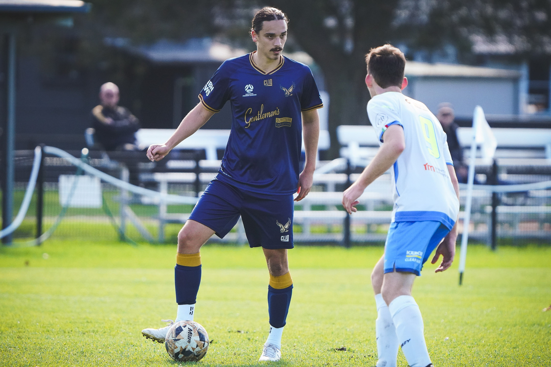 A soccer player in a dark blue jersey with the number 4 controls the ball while facing an opponent in a white jersey on a grassy field.