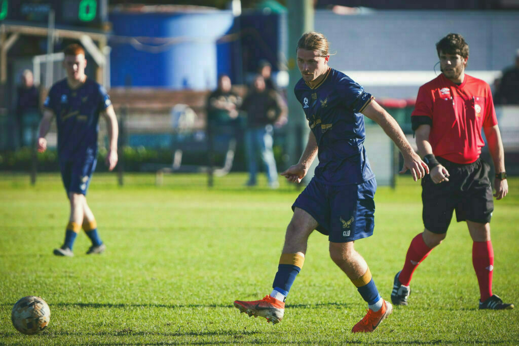 A soccer player in a blue uniform dribbles the ball while a referee in red watches, with another player in blue in the background.