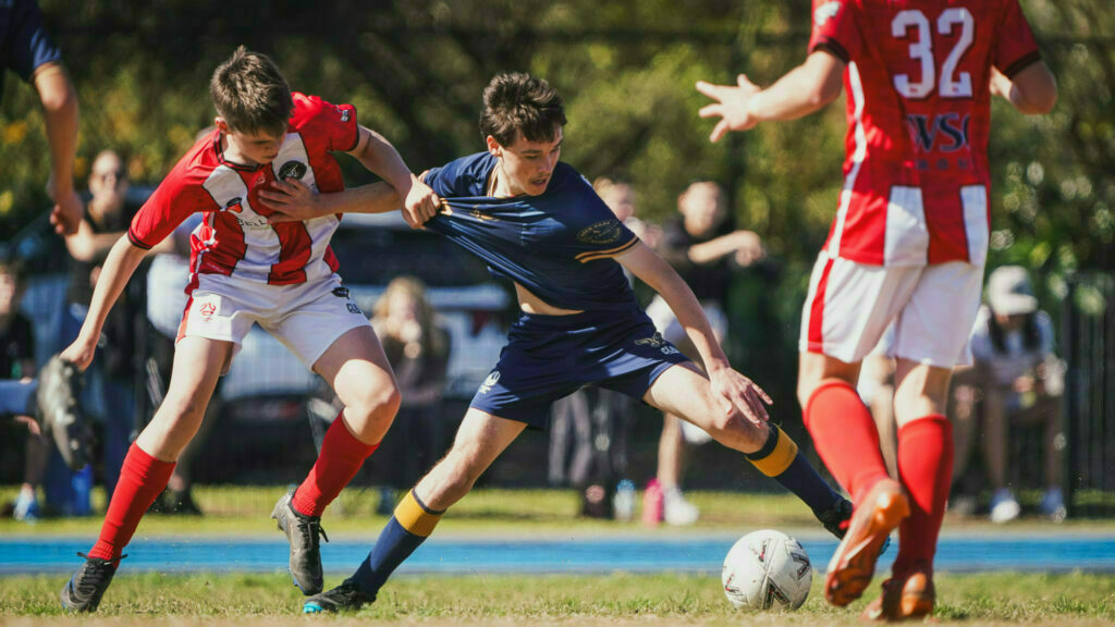 Two young soccer players in red and white jerseys are trying to block an opponent in a blue jersey who is attempting to gain control of the ball.