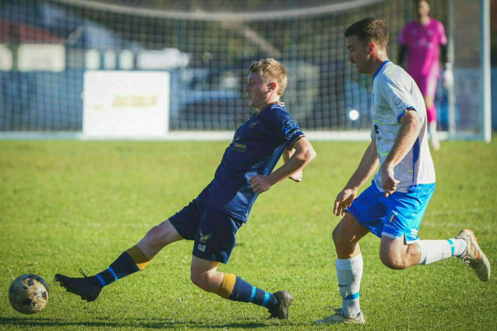 Two soccer players are engaged in action on the field, with one attempting to kick the ball while the other defends.