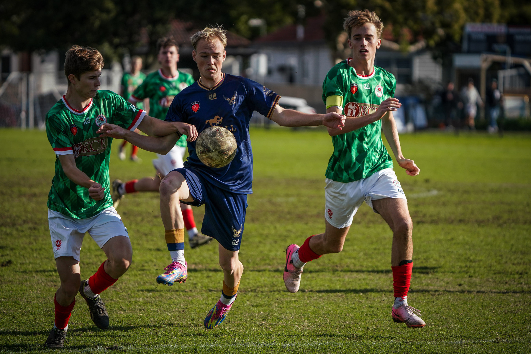 A soccer player in a blue jersey controls the ball with his foot while two opponents in green jerseys attempt to challenge him.