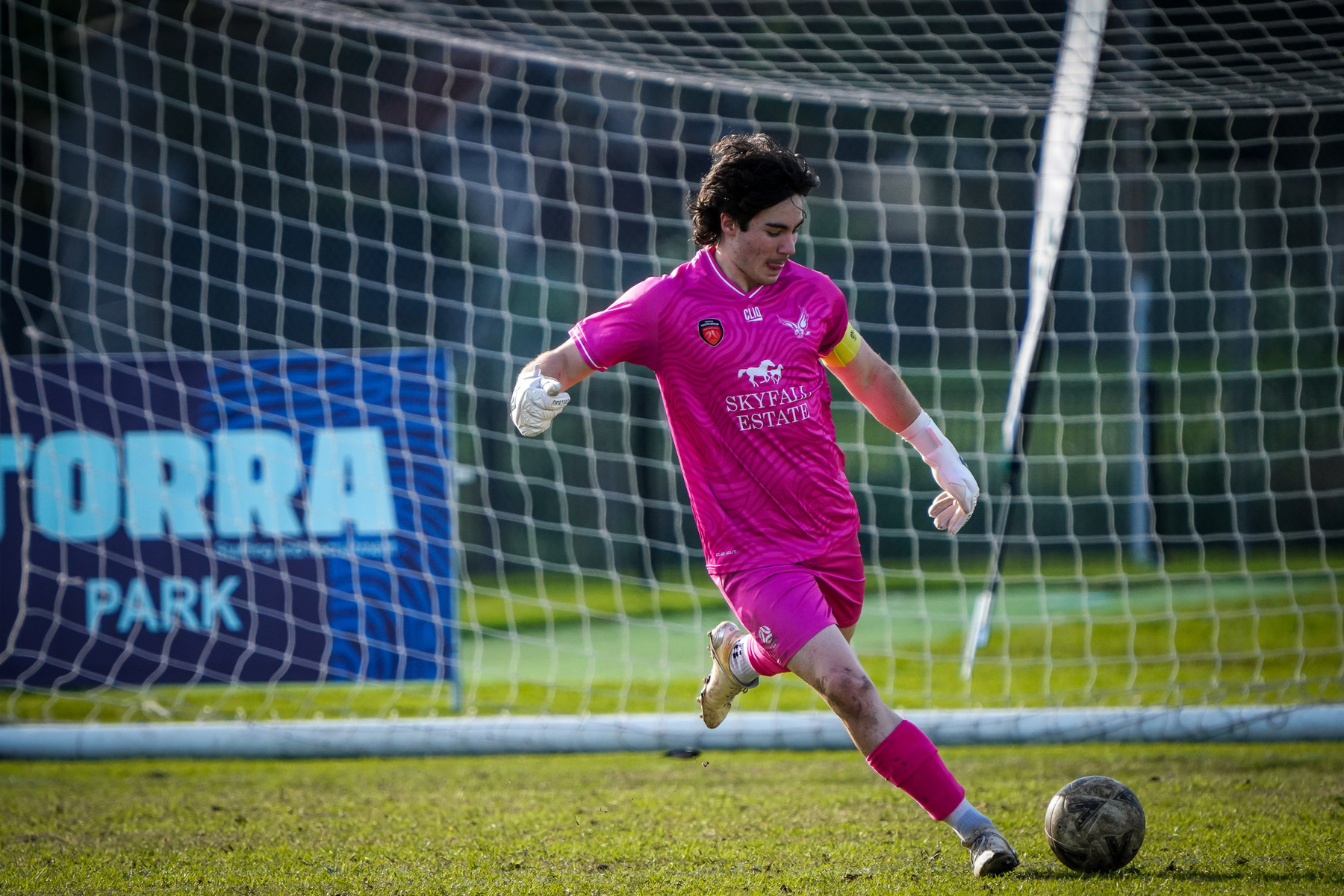 A soccer player in a pink uniform is kicking a ball on a grassy field in front of a goal net.