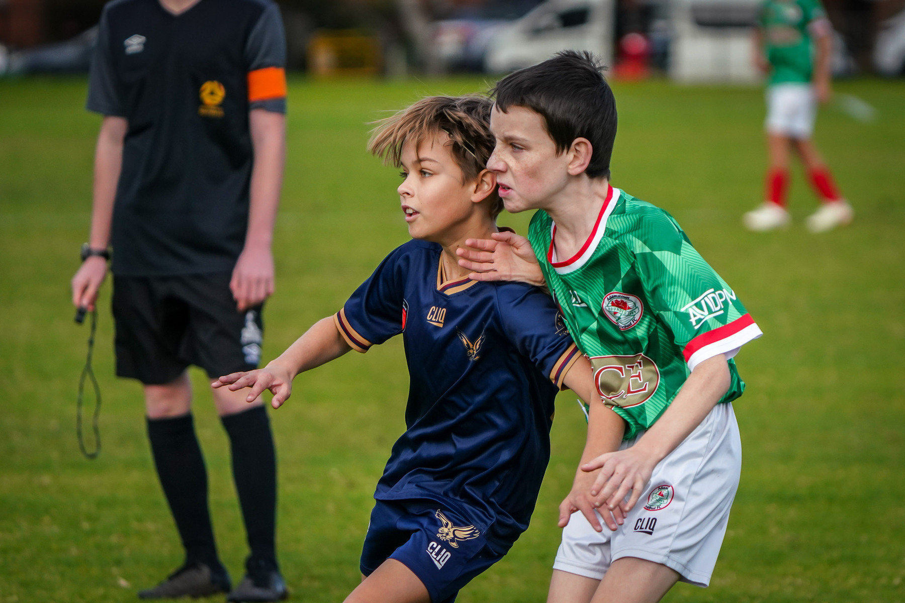 Two young soccer players compete for position on the field, with a referee in the background.