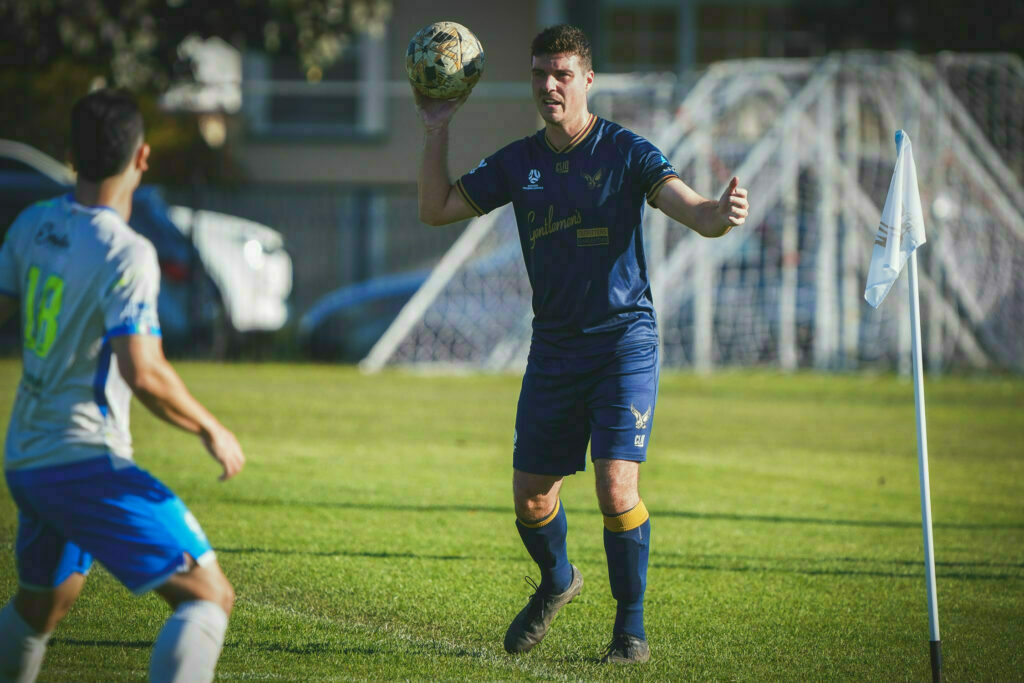 A soccer player wearing a blue uniform is about to throw a ball back into play near the corner flag, while an opposing player in a light gray and blue uniform approaches.