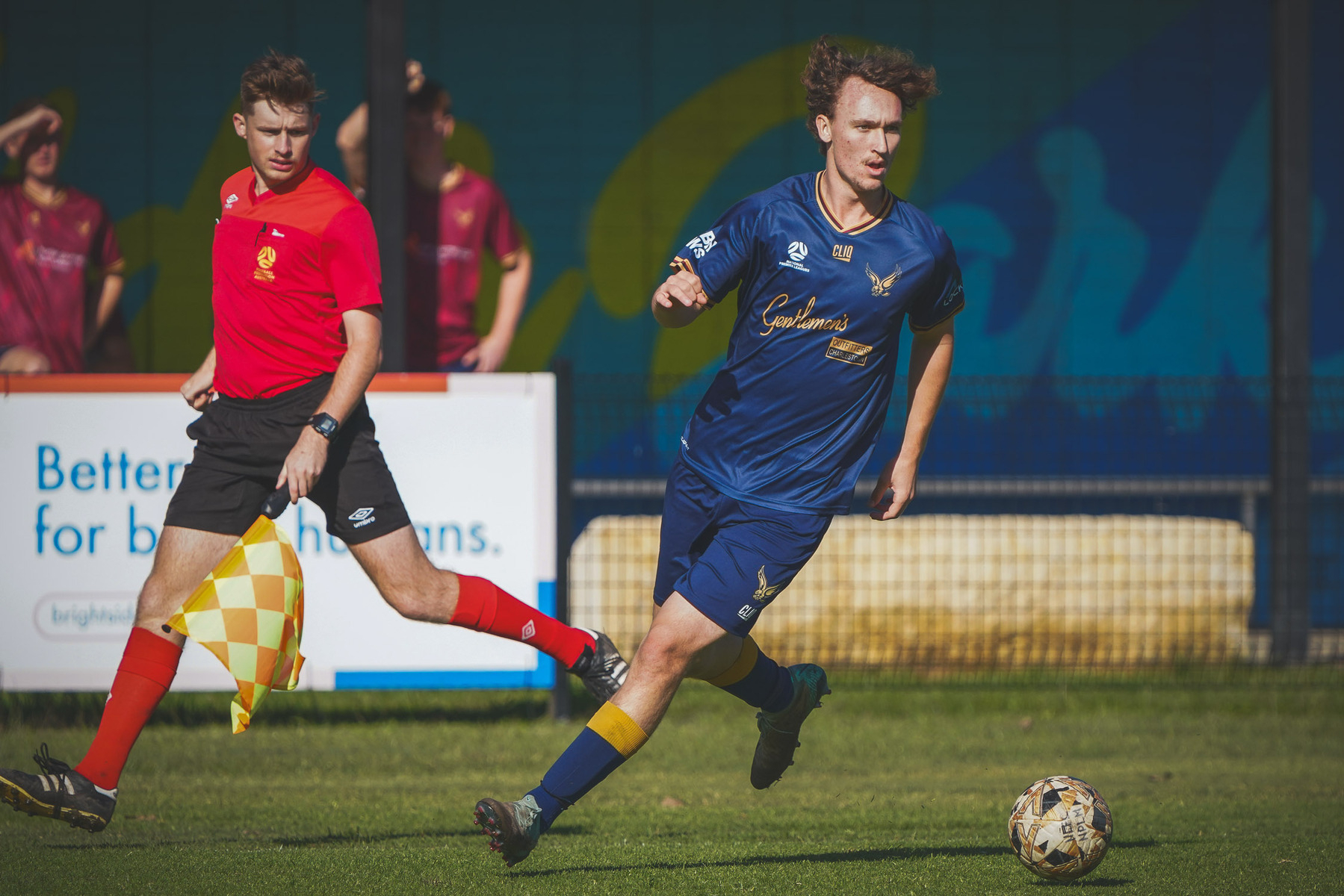 A soccer player in a blue uniform runs on the field while a referee follows behind.