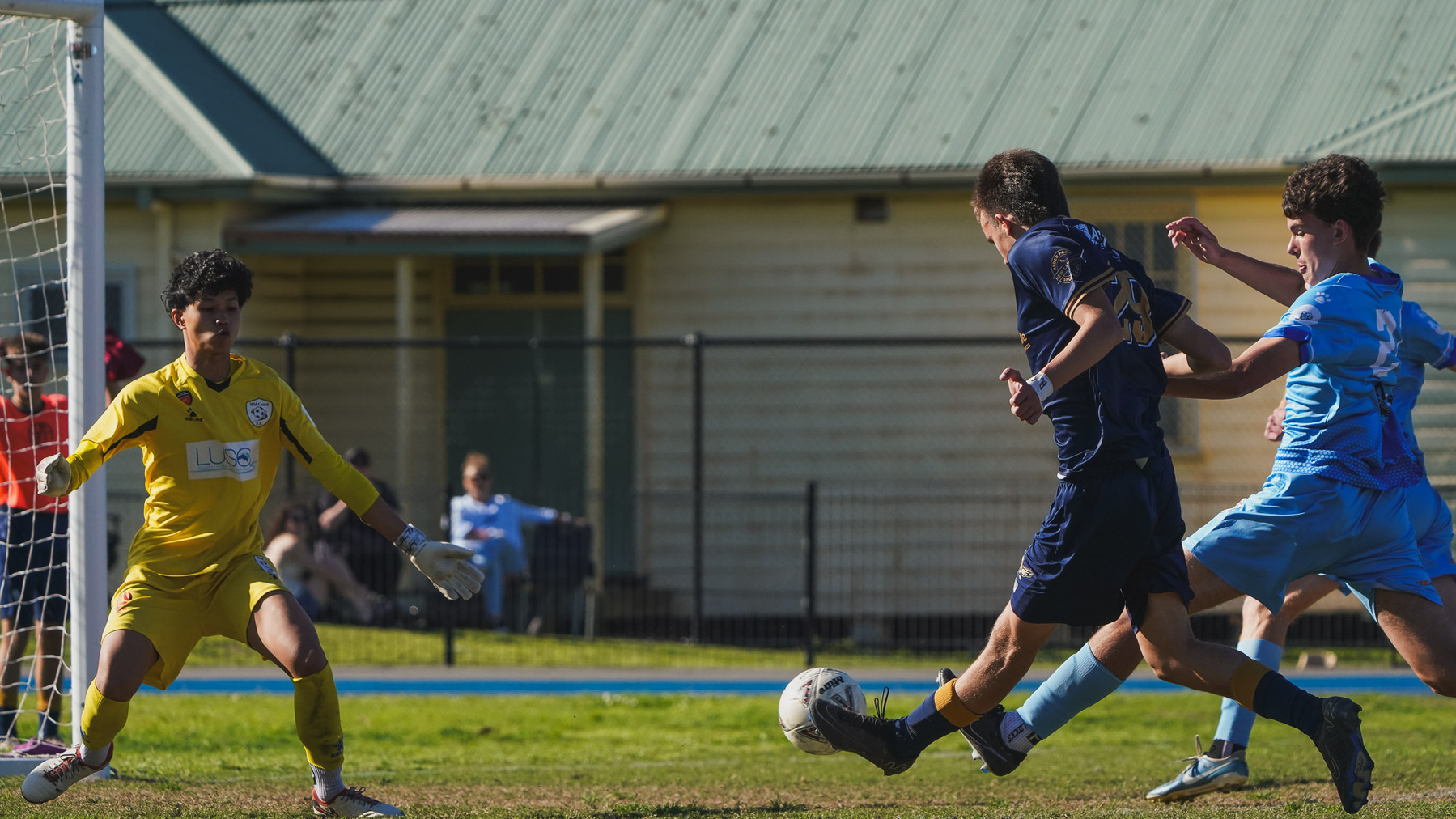 A soccer match features two players in light blue trying to defend against a player in navy as a goalkeeper in yellow awaits the action near the goal.