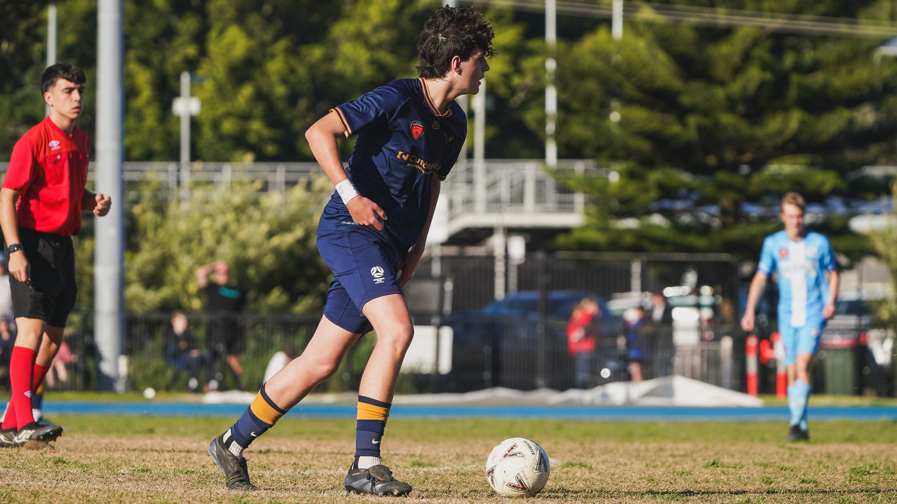 A soccer player in a navy uniform is on a grassy field, ready to move the ball as other players and a referee are nearby.