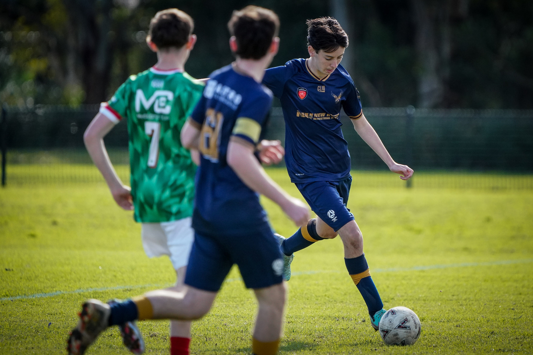 A soccer player in a blue jersey prepares to kick the ball while two opponents approach in green jerseys.