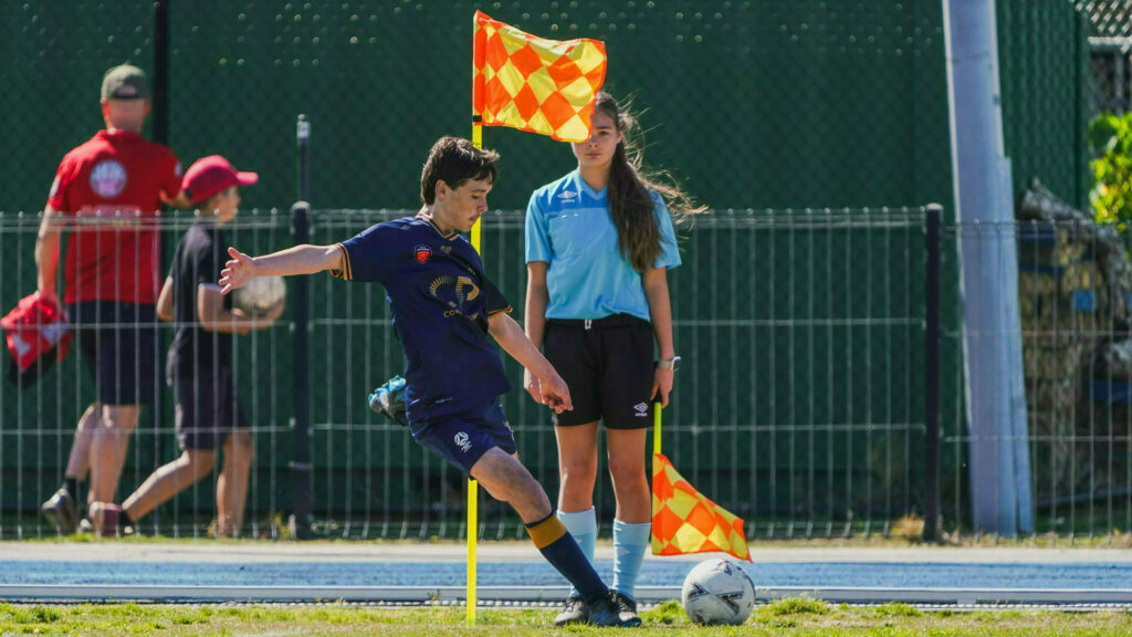 A young soccer player kicks the ball near the corner flag during a game, with a referee and spectators in the background.