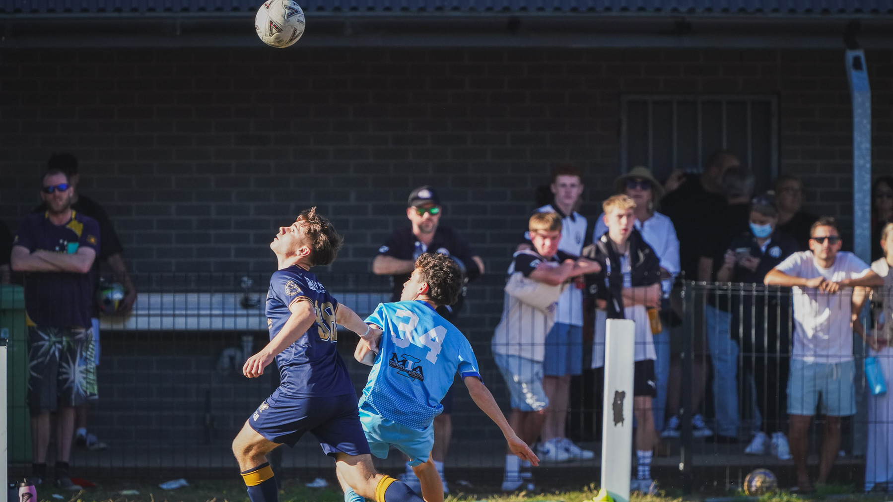 Two soccer players are competing for a ball in the air while a group of spectators watches from the sidelines.