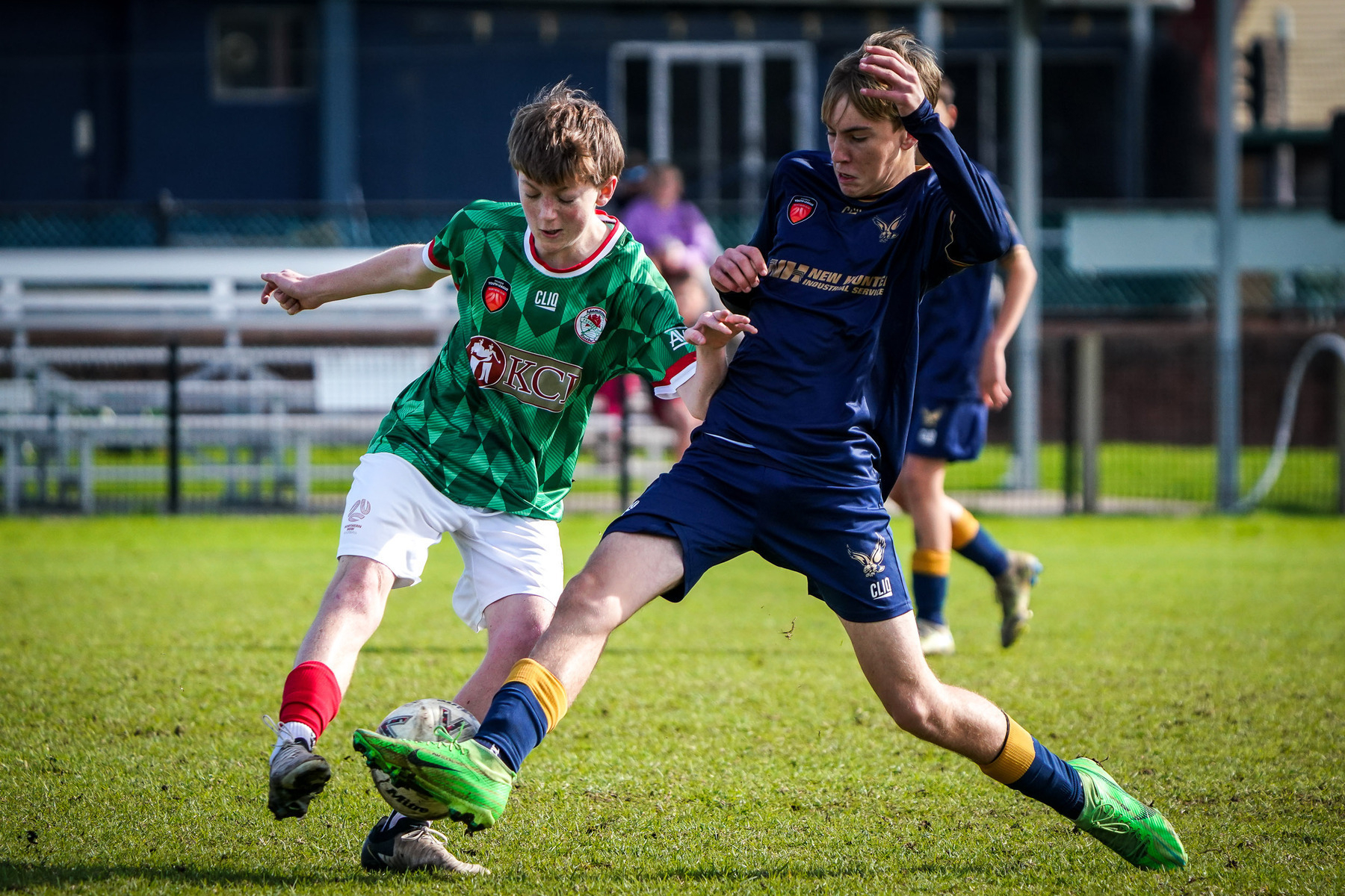 Two soccer players in green and blue uniforms compete for the ball on a grassy field.