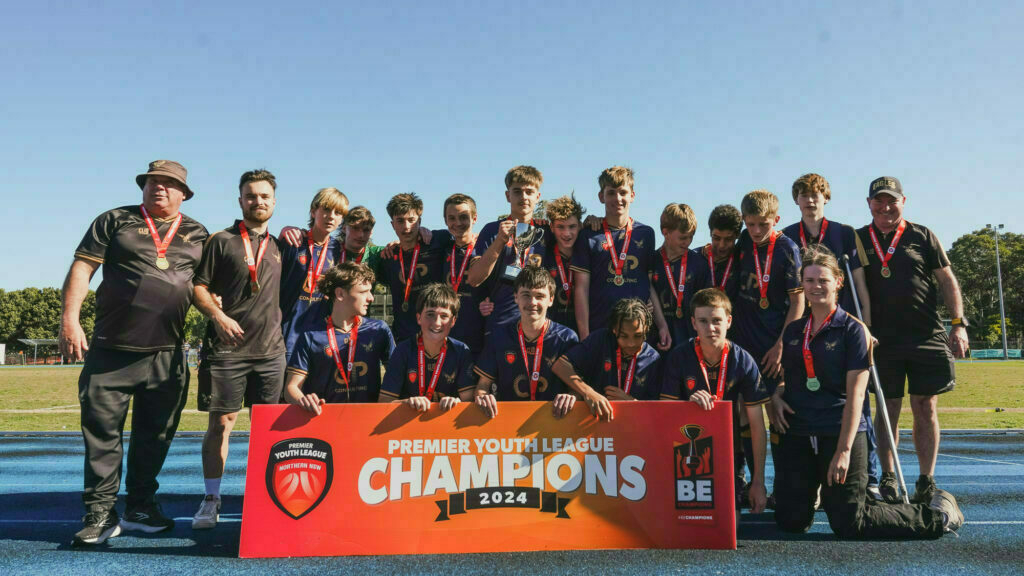 A soccer team poses with a Premier Youth League Champions 2024 banner and medals on a field.