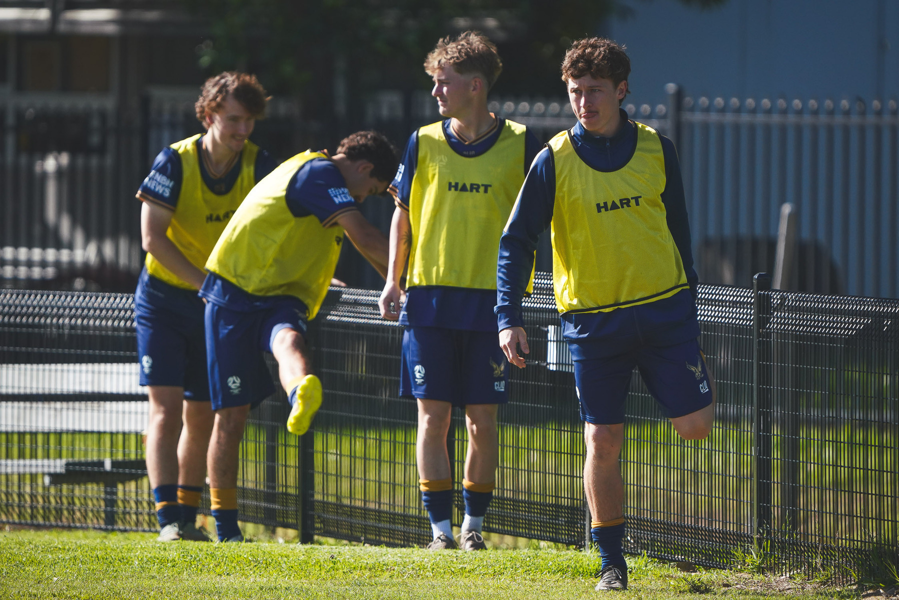 A group of young soccer players wearing yellow bibs is seen stretching and warming up near a fence.