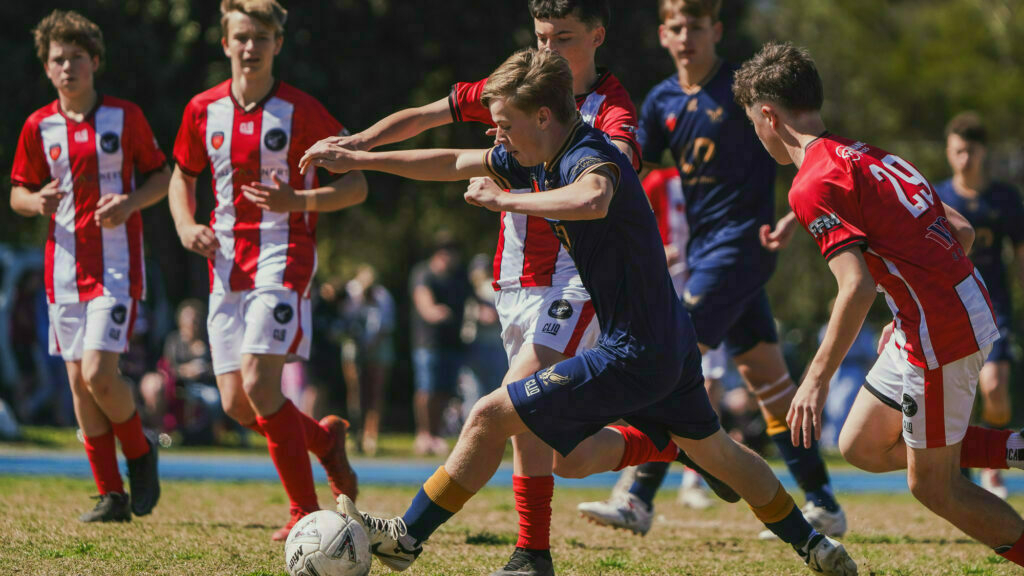 A group of young soccer players wearing different colored uniforms are actively engaged in a match, with one player in a dark uniform dribbling the ball while others in red-striped uniforms attempt to intercept.