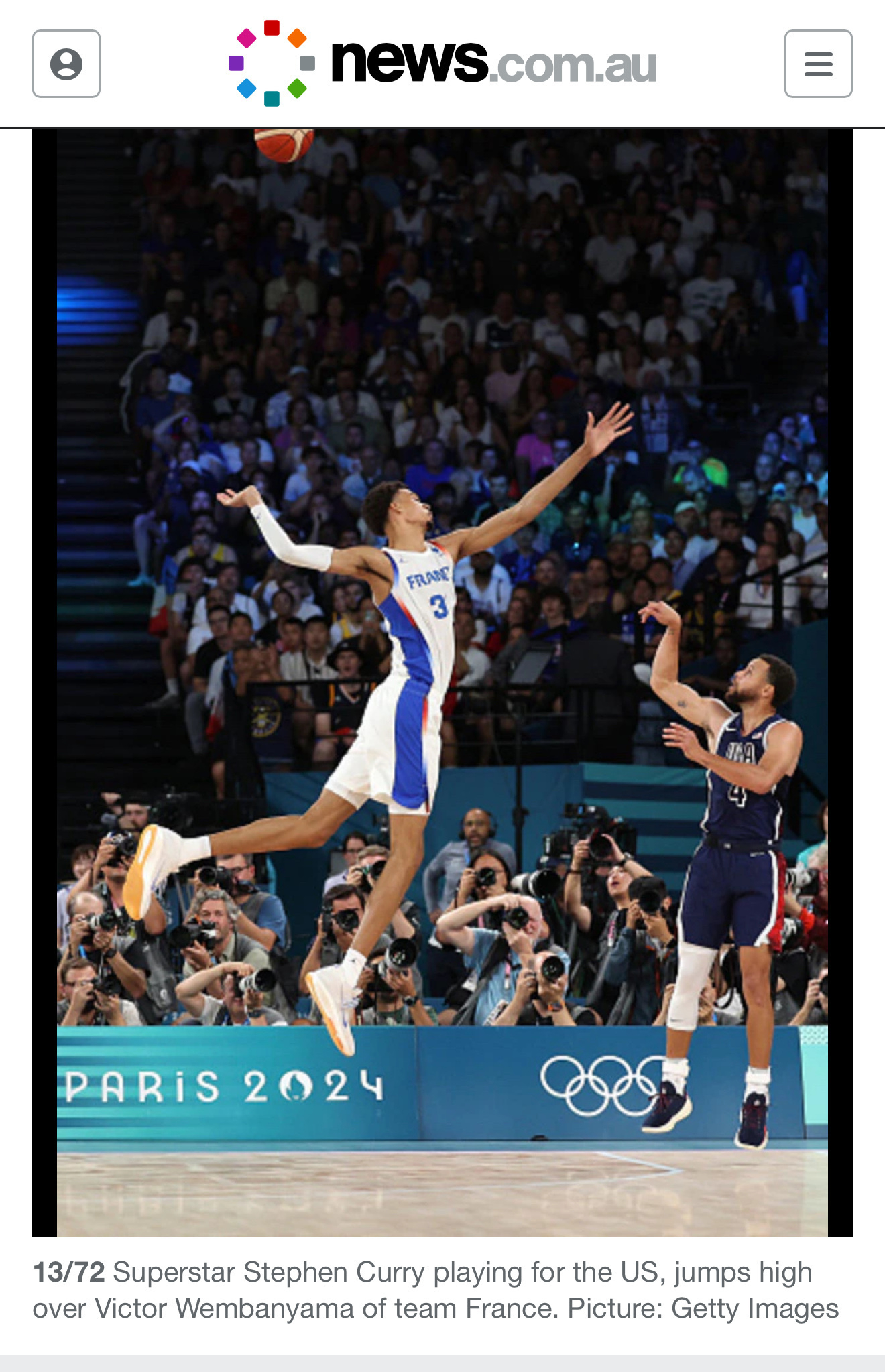 A basketball player in a blue and white France jersey leaps high to block a shot from an opposing player in a dark USA jersey during a game, with spectators watching in the background.