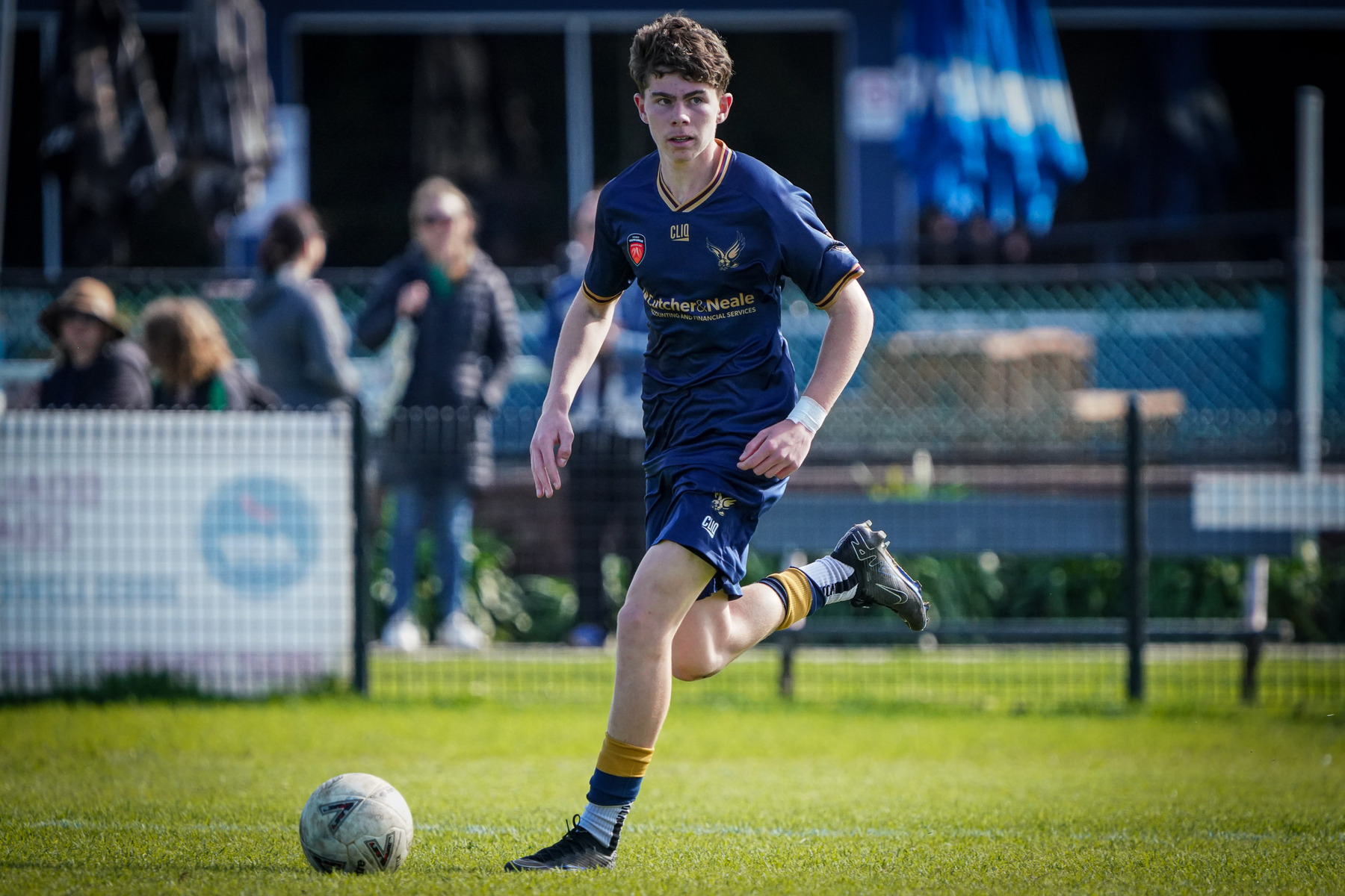 A young soccer player in a blue and gold uniform is running on a grassy field while dribbling a ball.