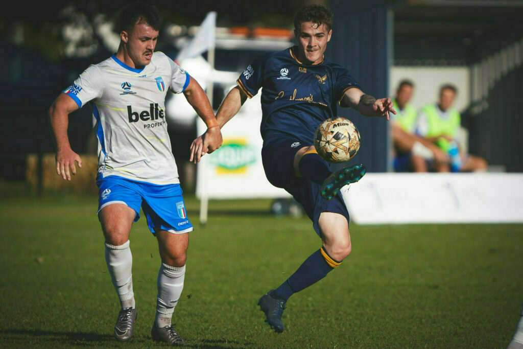 Two soccer players are competing for the ball on a grassy field, with one player attempting to intercept the ball mid-air.