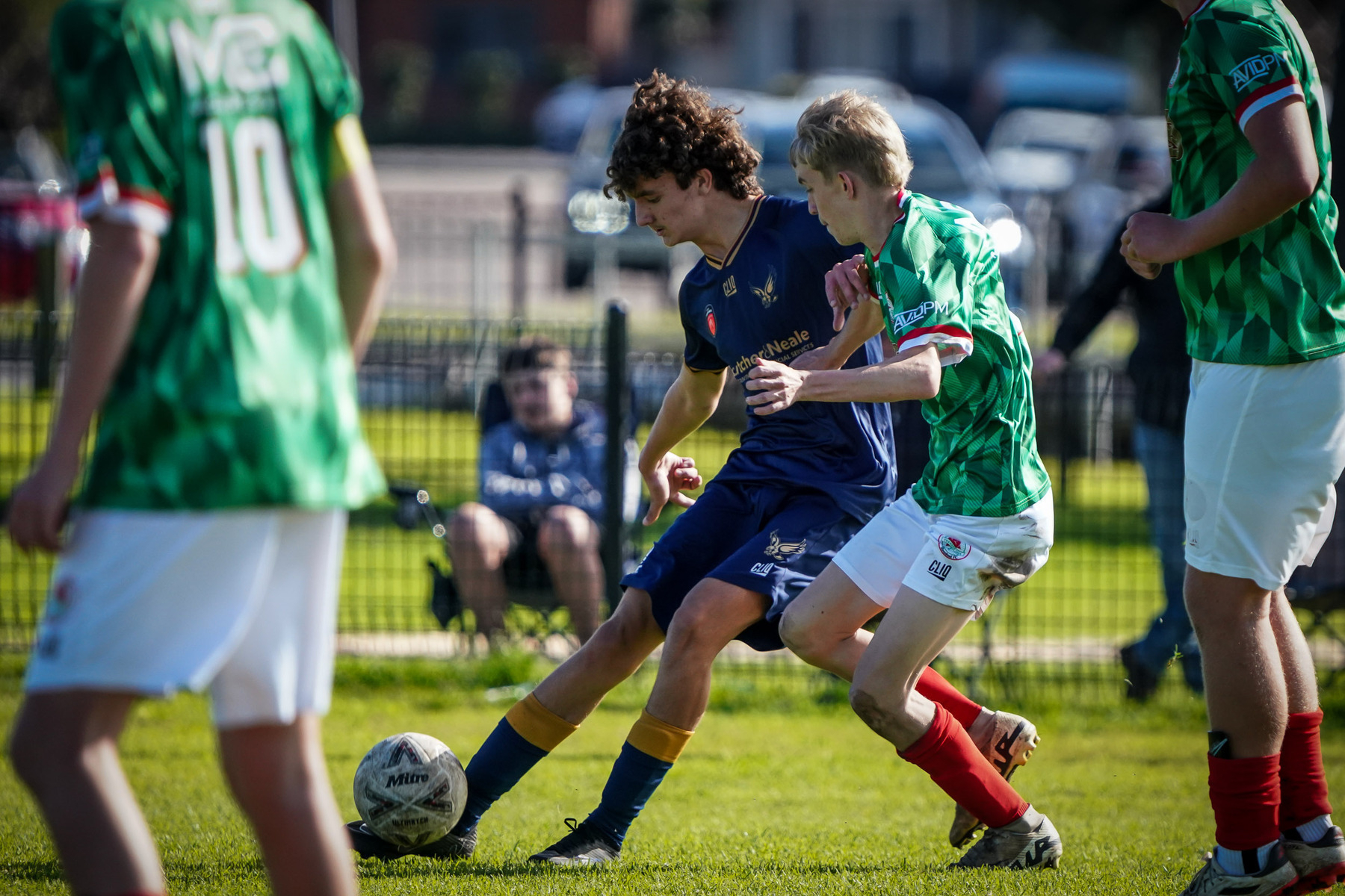 Two soccer players are competing for the ball on a grassy field, with teammates and spectators in the background.