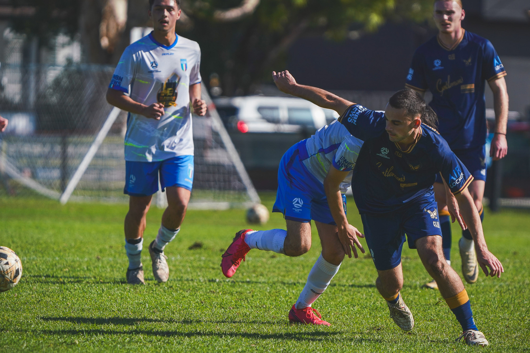 Two soccer players are in action on the field during a game, with one wearing a blue jersey and the other in a white and blue jersey, while two other players look on.