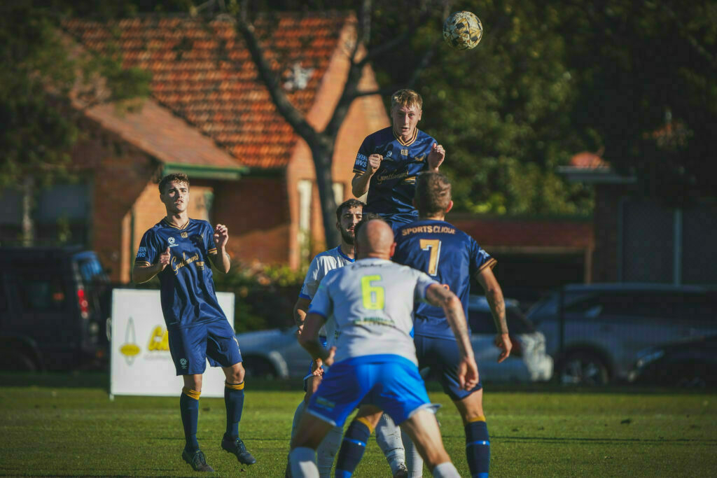 A group of soccer players in blue and white jerseys are actively competing for a ball in mid-air on a grassy field.