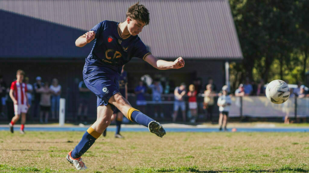 A soccer player in a navy blue uniform is kicking a ball on a field during a game, with spectators in the background.