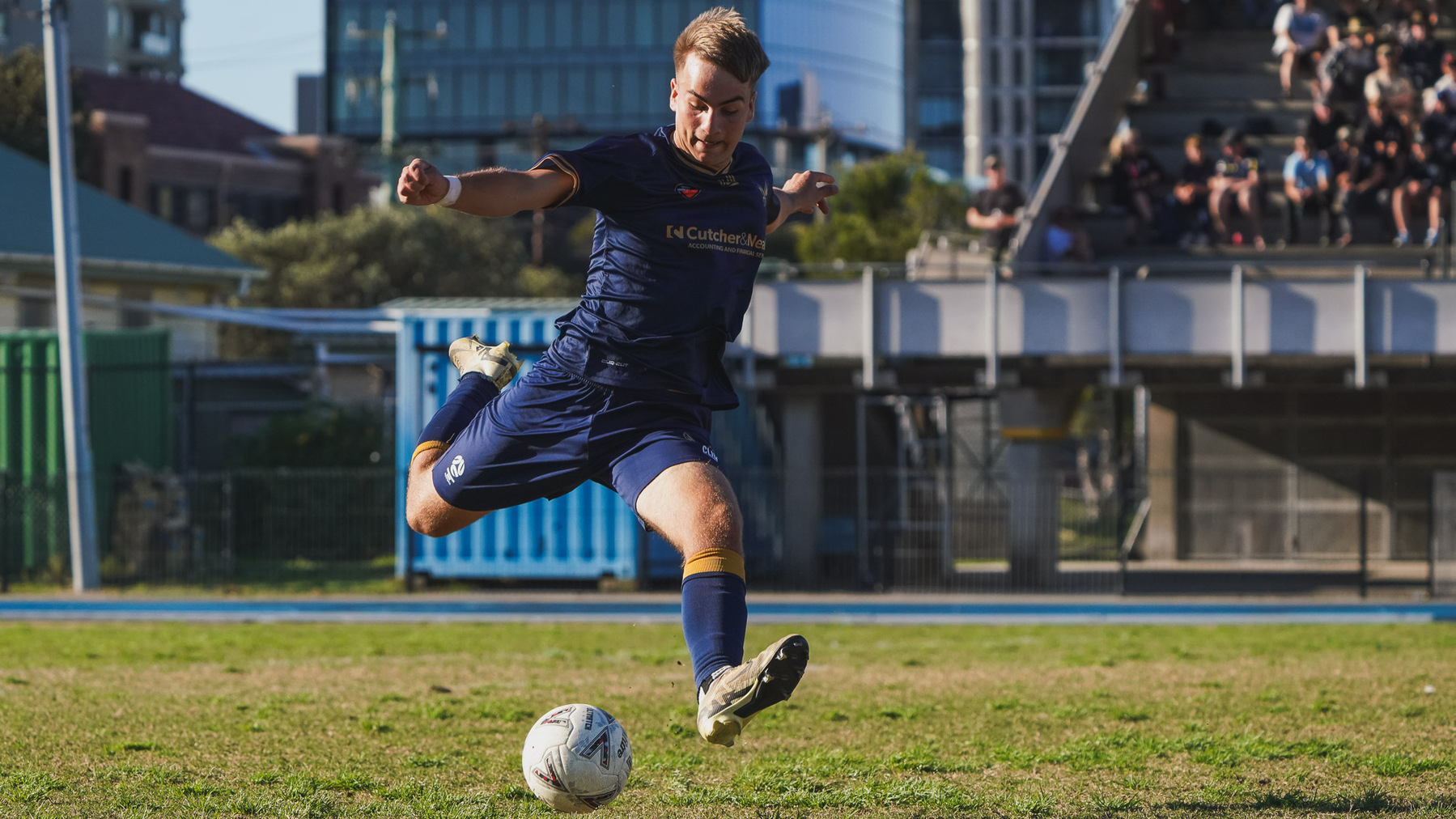 A soccer player in a blue uniform is kicking a ball on a grassy field with spectators in the background.