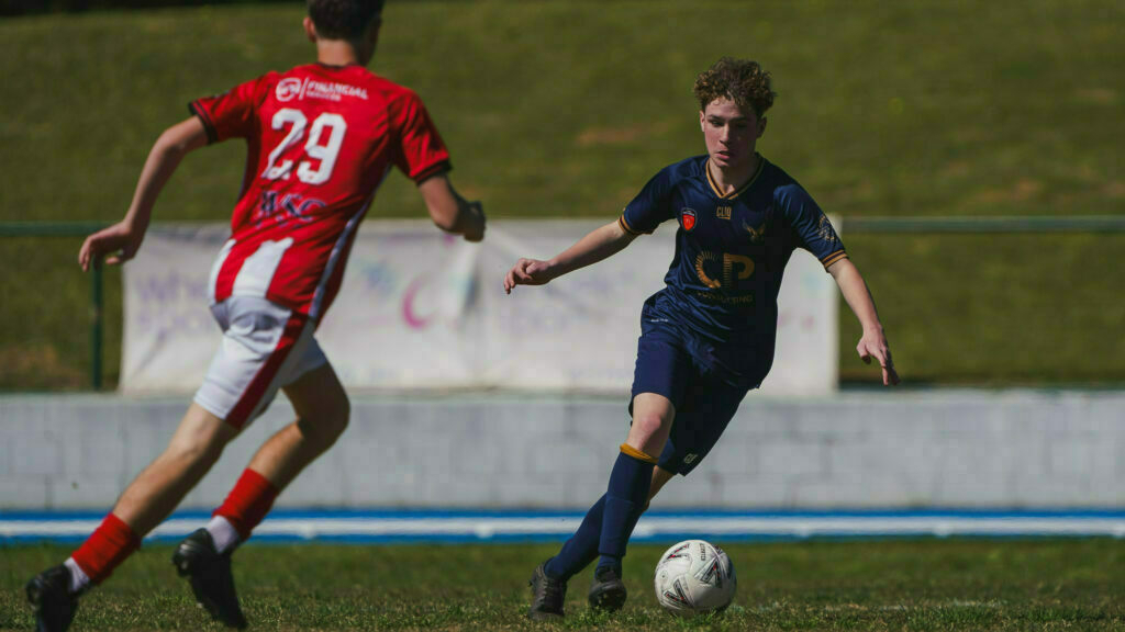 Two soccer players compete for the ball on a grassy field, with one in a red and white uniform and the other in a navy blue uniform.