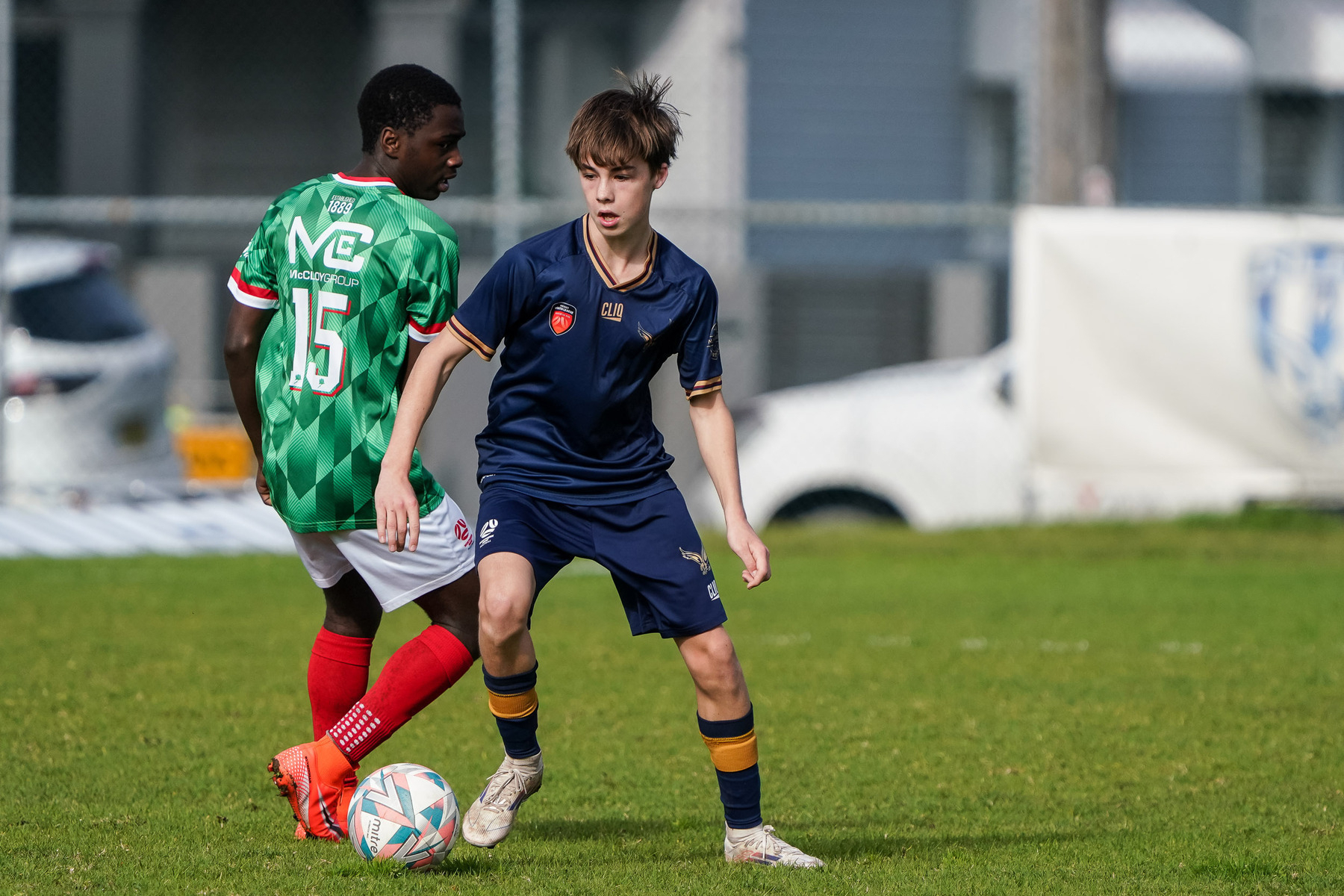 Two young soccer players are competing for the ball on a grassy field, one wearing a green and red uniform and the other in a navy blue uniform.