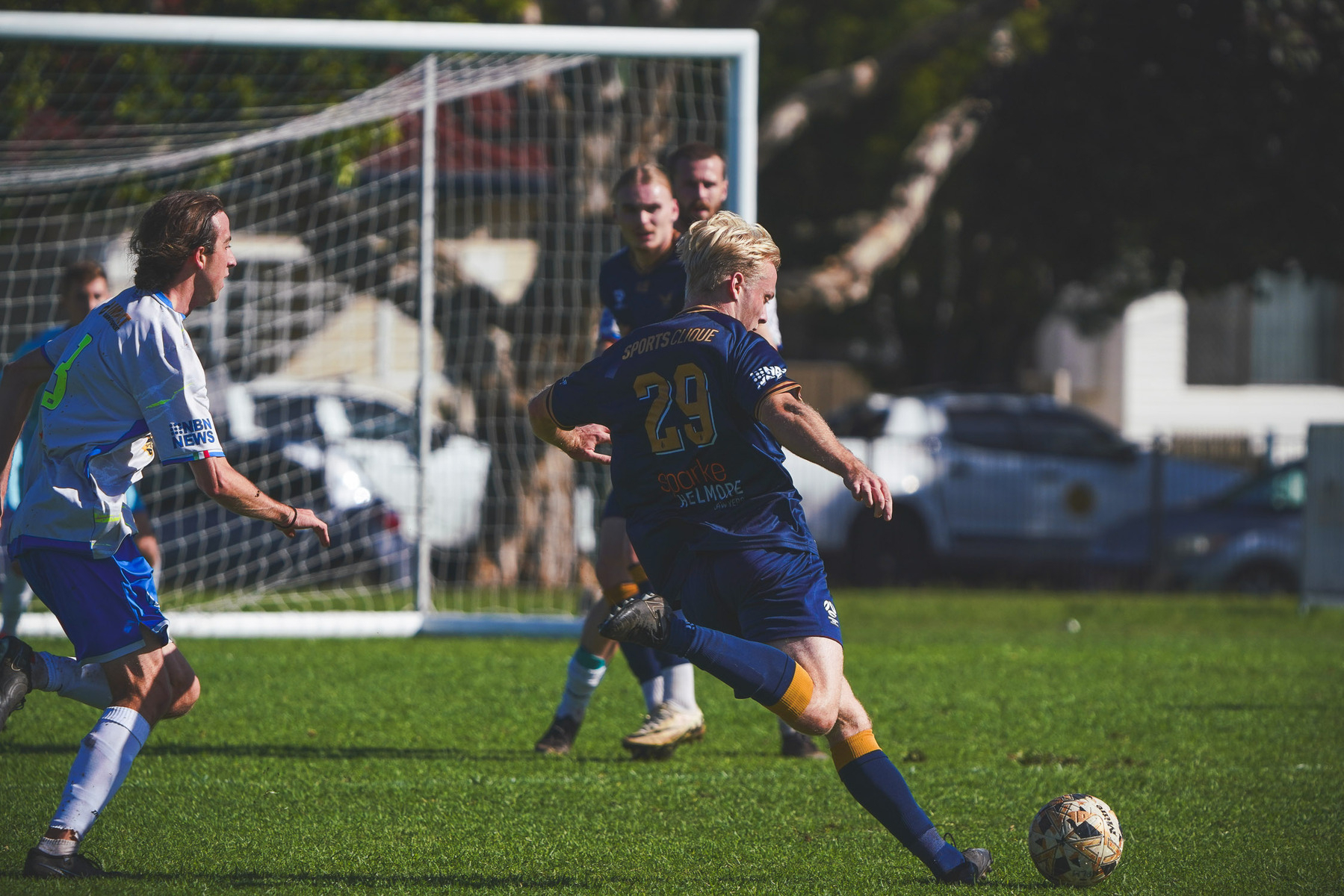 A soccer player in a blue jersey, numbered 29, kicks the ball on a green field while opponents approach, and a goal with netting looms in the background.