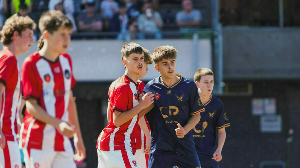 A group of boys in red and blue soccer uniforms are focused on the game on a sunny day, with spectators in the background.