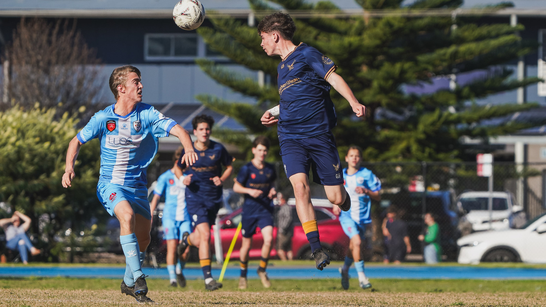 Two soccer players, one in light blue and one in dark blue, are competing for a ball in mid-air during a match.