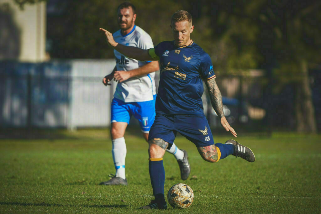 A soccer player in a blue jersey is about to kick a ball during a match, with another player in a white jersey in the background.