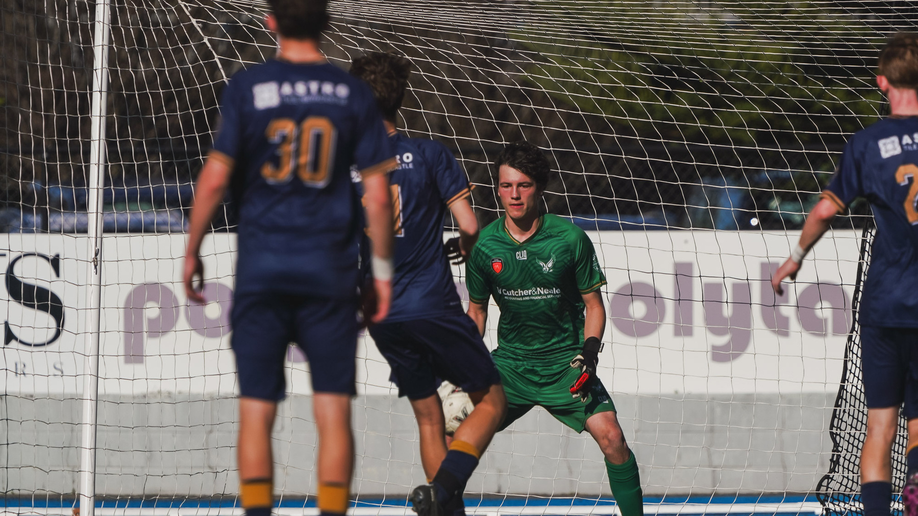 A soccer goalkeeper in green gear is focused on the ball as opposing players in blue uniforms move towards the goal.