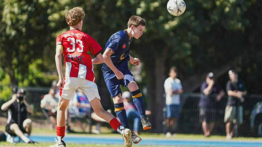 Two soccer players in action during a game, with one jumping to head the ball.