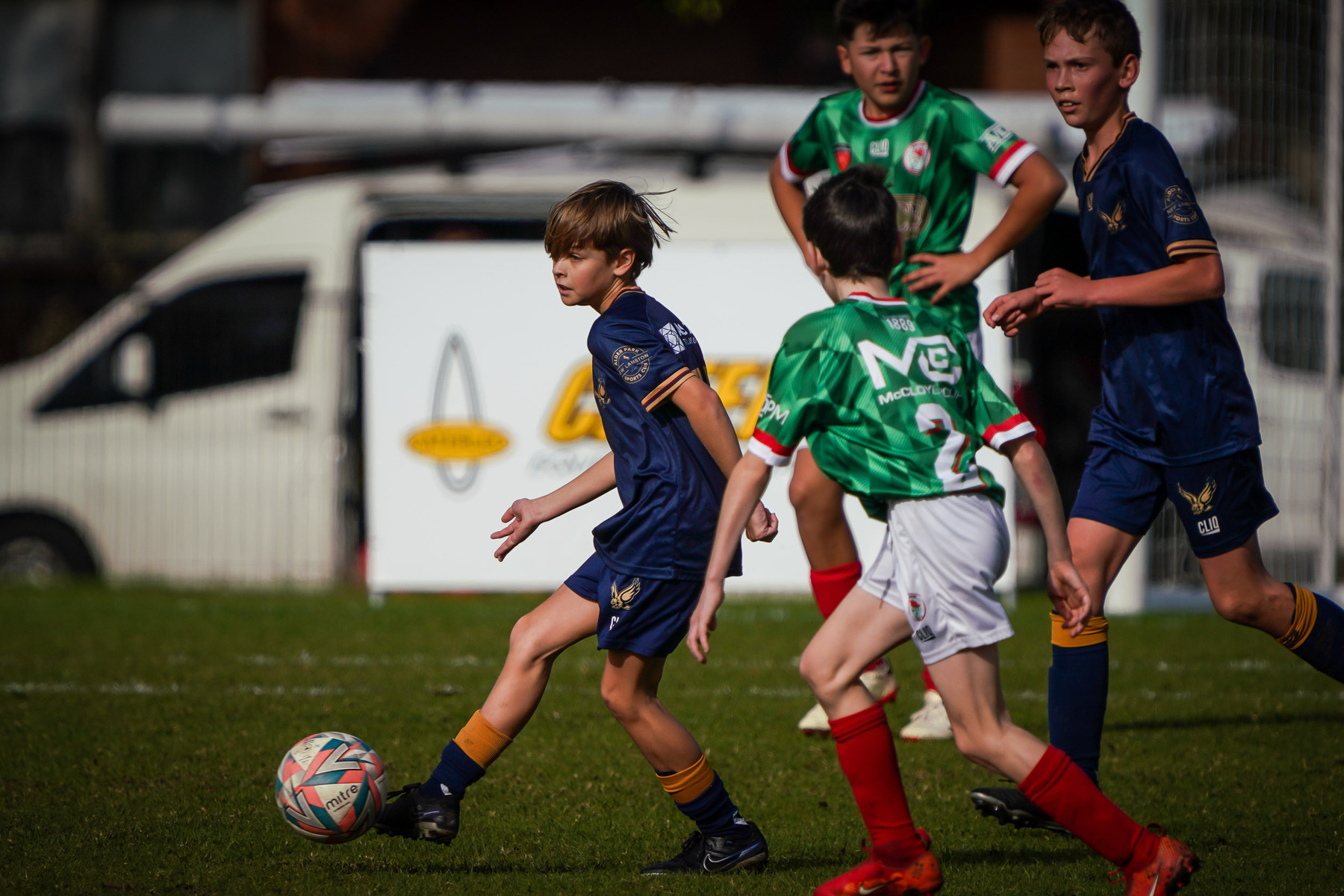 Several young soccer players in blue and green jerseys are actively playing on a grassy field, with one player in focus controlling the ball.