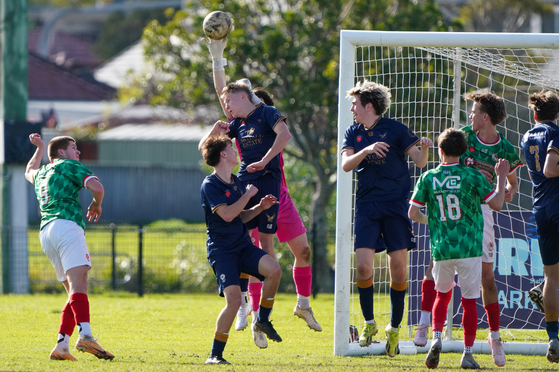 Several soccer players in green and blue jerseys are attempting to head or block the ball near the goal during a match.