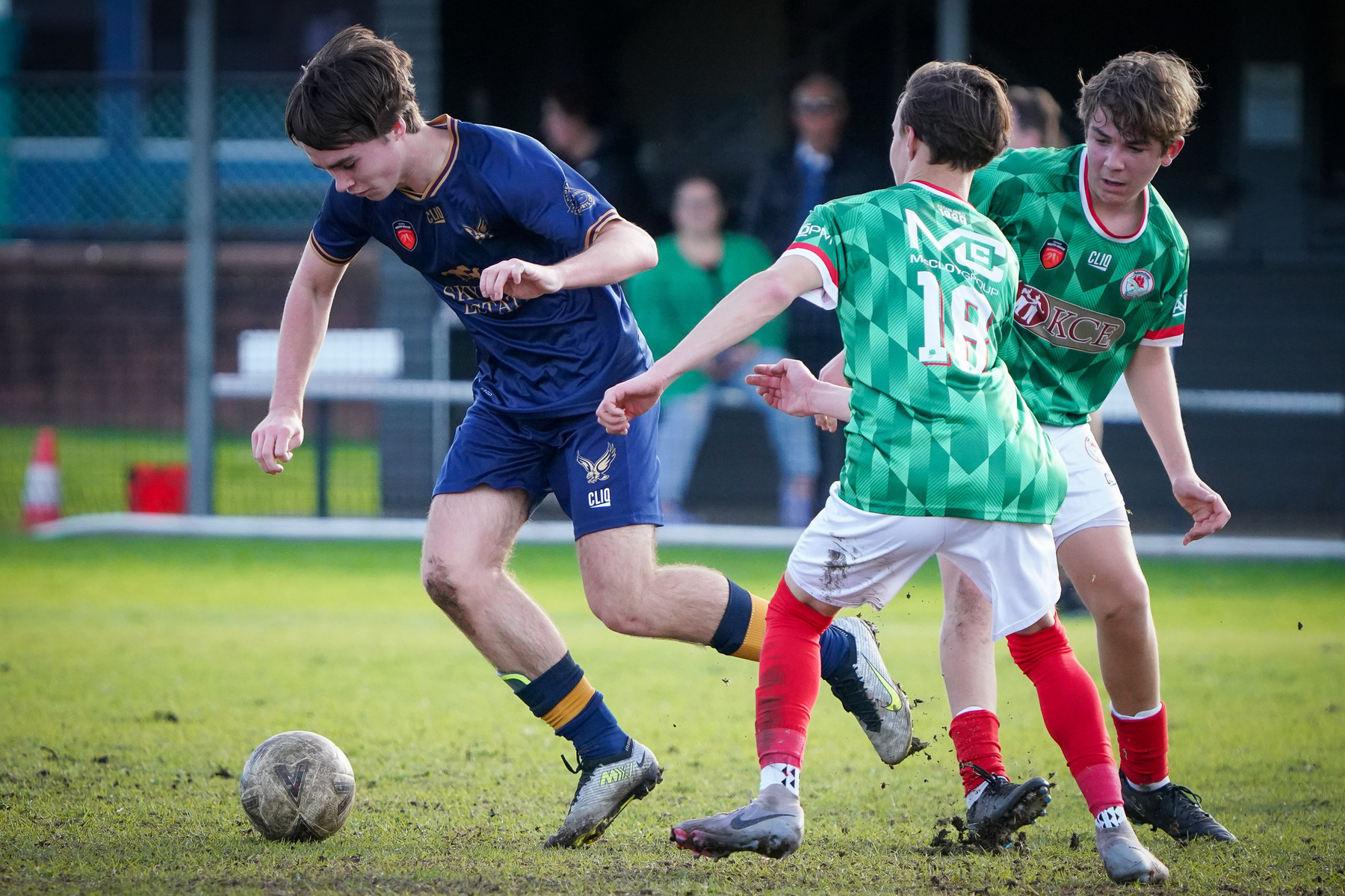 A young soccer player in a blue uniform dribbles the ball past two opposing players in green jerseys on a grassy field.