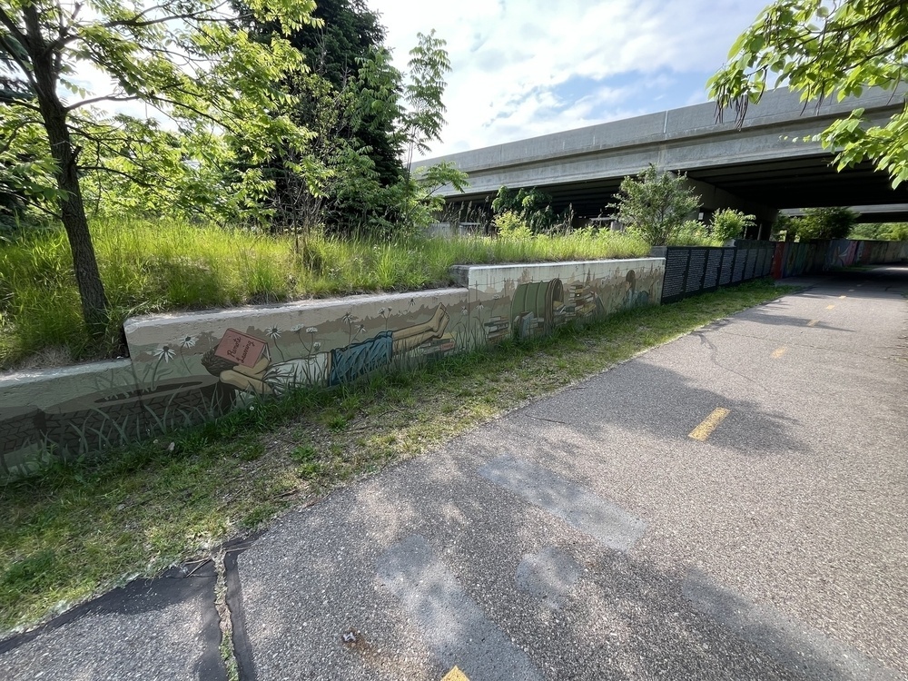 A pathway runs alongside a mural-covered wall under an overpass, surrounded by greenery and trees.