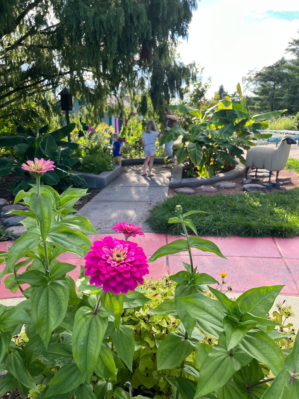 Bright pink flowers are prominently in the foreground of a garden scene where children and an adult walk along a stone pathway surrounded by lush greenery and a decorative sheep statue.