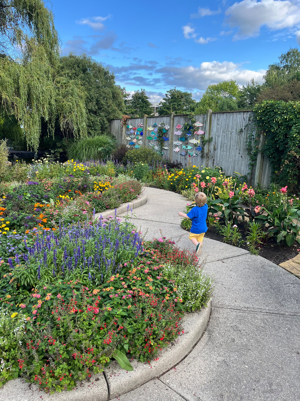 A young child is walking along a curved garden path surrounded by colorful flowers and lush greenery under a partly cloudy blue sky.