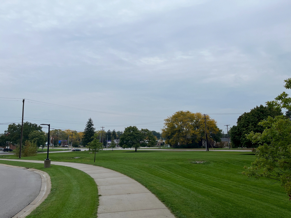 A curved sidewalk runs alongside a well-maintained grassy area with scattered trees under a cloudy sky.
