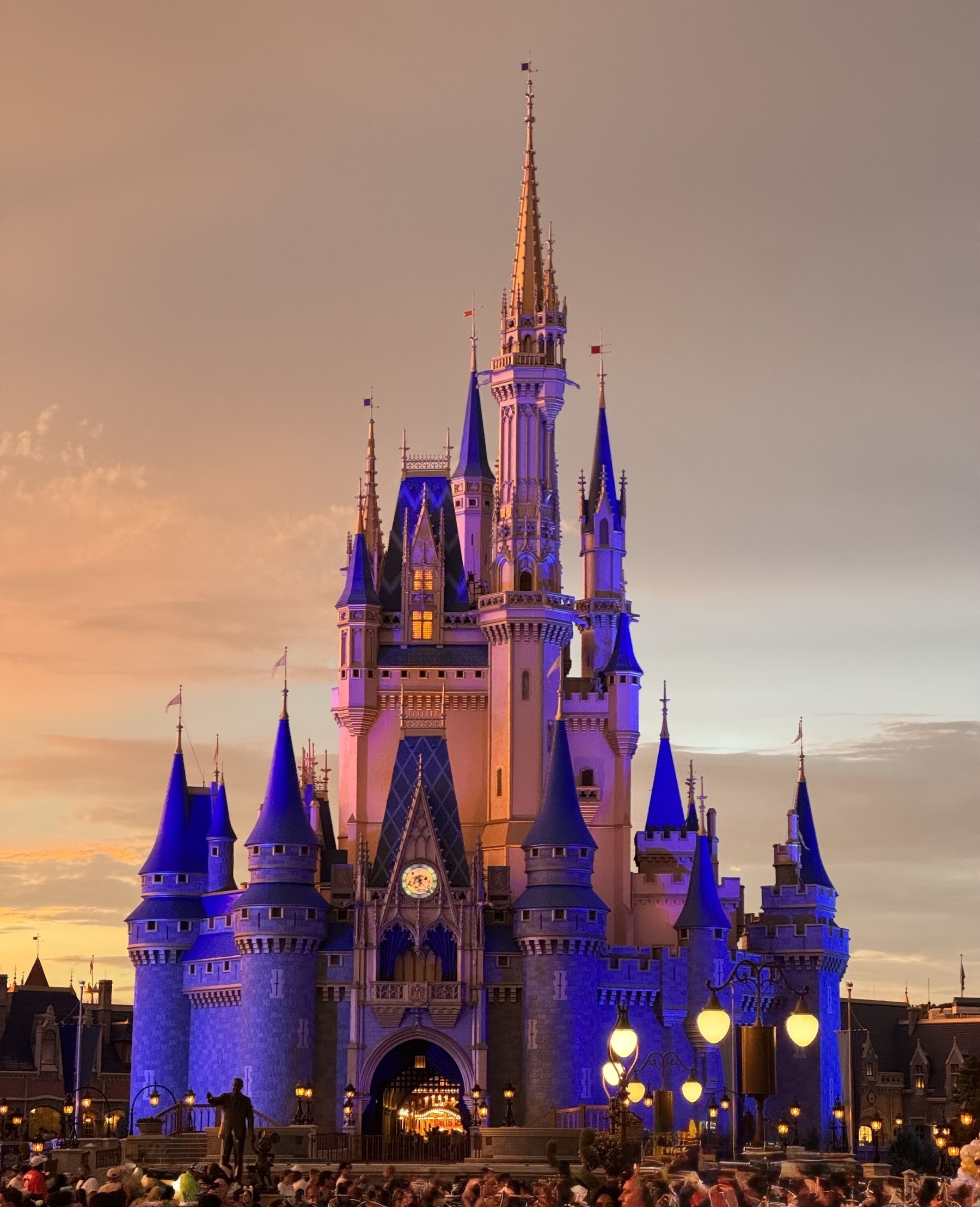 A brightly illuminated Cinderella Castle stands against a colorful sunset sky, surrounded by a crowd of people.