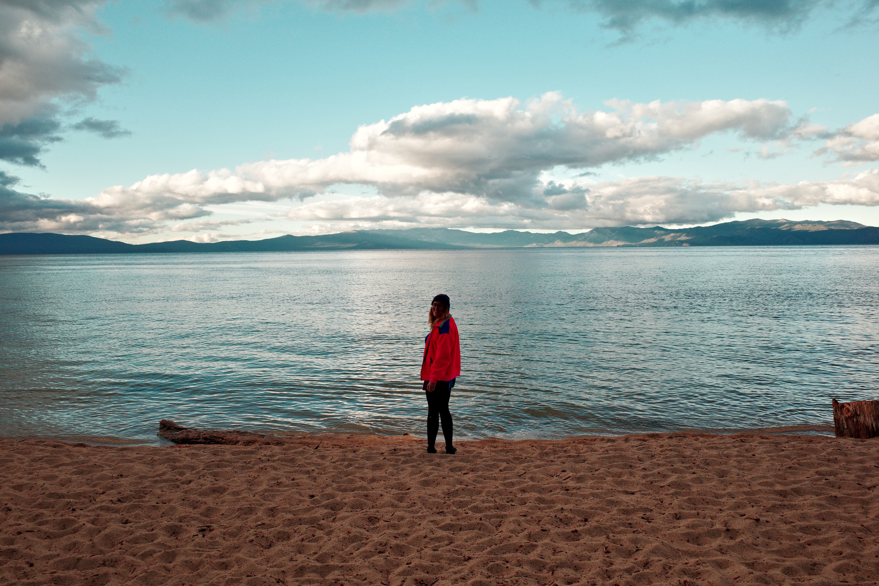 A person wearing a red jacket stands alone on a sandy beach looking out over a calm body of water with mountains and clouds in the distance.